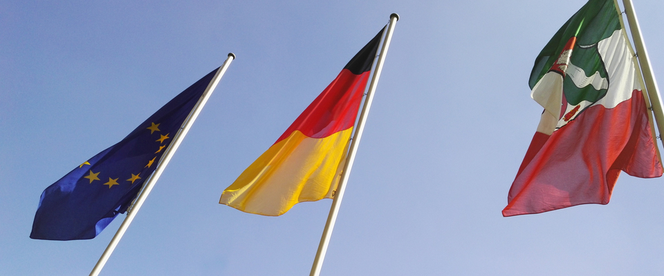 Drei wehende Flaggen vor blauem Himmel, von links nach rechts:
Europa-Flagge
Deutschland-Flagge
NRW-Flagge